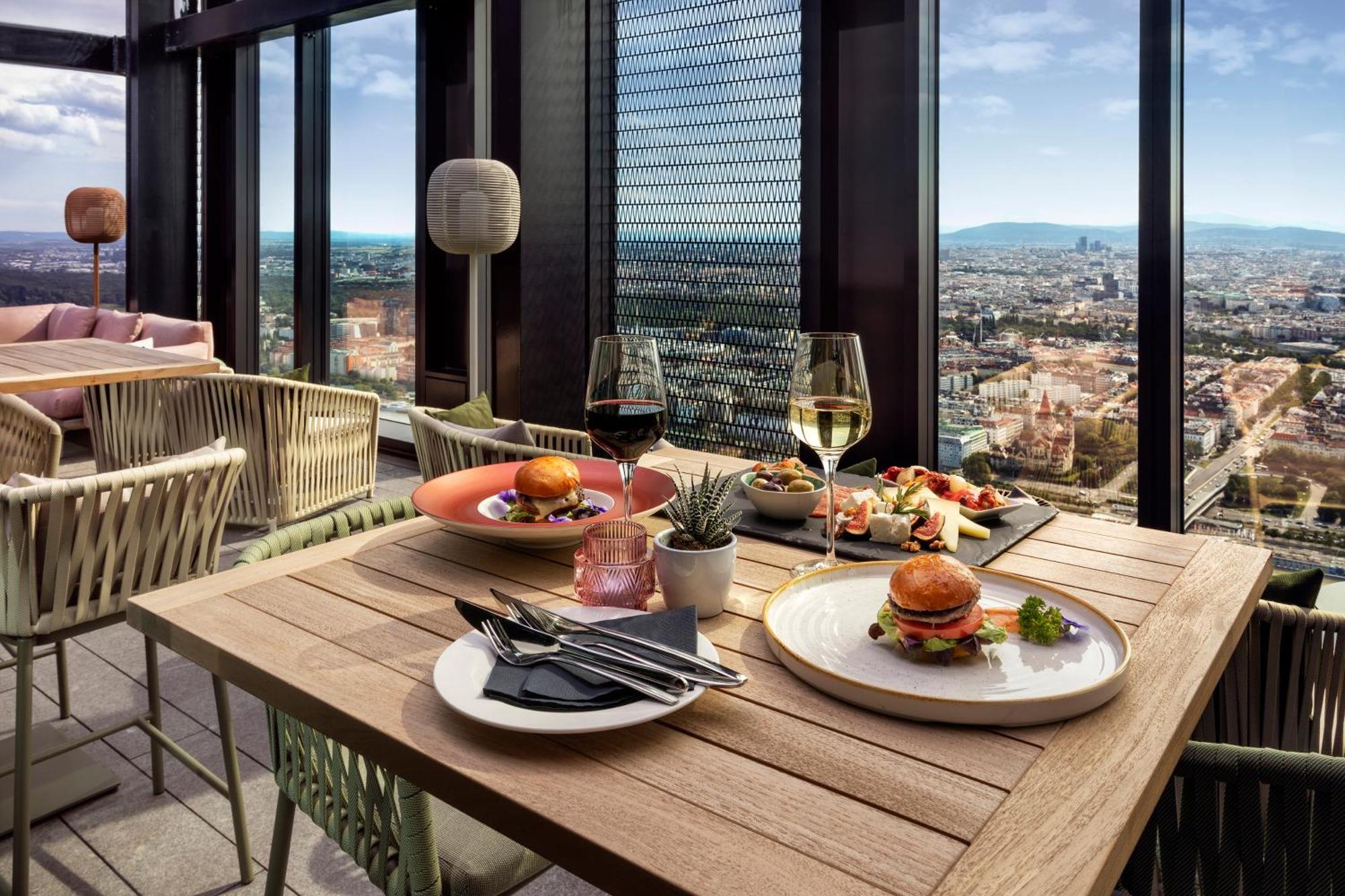 เมเลีย เวียนนา Hotel ภายนอก รูปภาพ The photo shows a stylish dining setup on a high rooftop or terrace, with a panoramic view of a city skyline in the background. 

There are two tables made of wood; one features two glasses of white wine and an assortment of dishes, including a plate