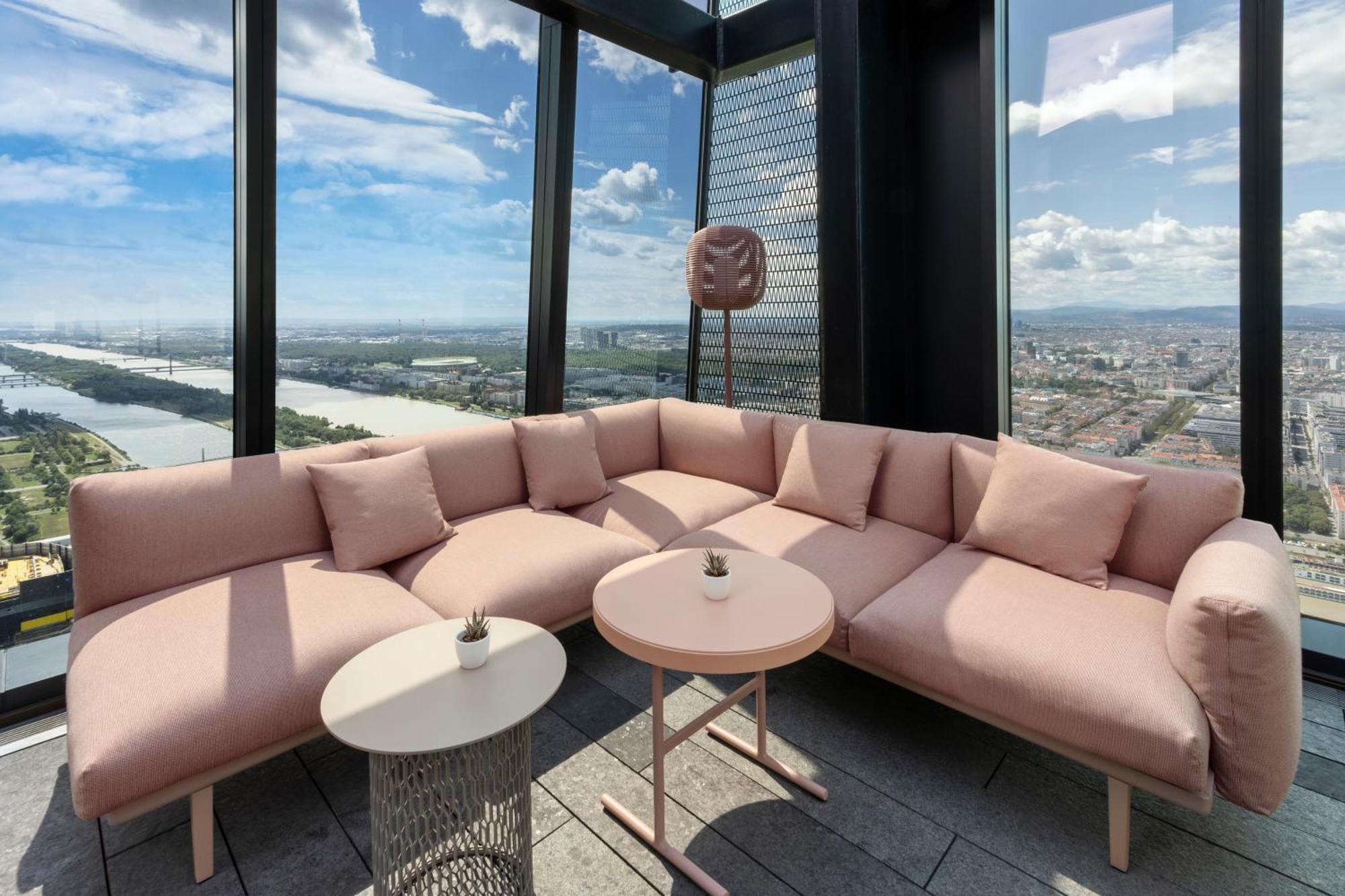 เมเลีย เวียนนา Hotel ภายนอก รูปภาพ The photo shows a modern lounge area with a large, L-shaped pink couch adorned with several soft cushions. In front of the couch, there are two tables: one round and one with a more intricate design. The space is located in a high-rise building, offe